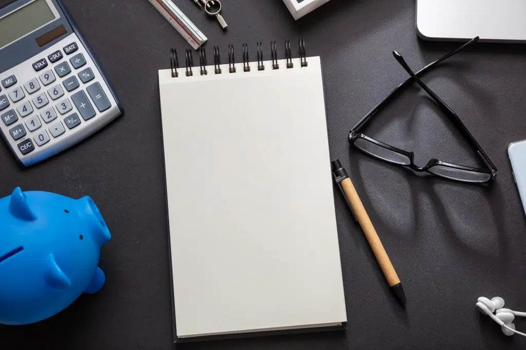 Mesa com itens espalhados, cofrinho, agenda, caneta, óculos, calculadora, compasso e escalimetro